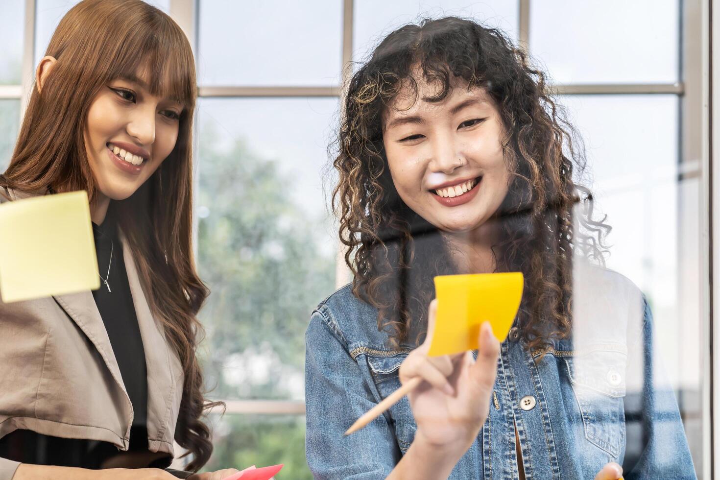 Asian creative business women are writing note on the text sheet after thinking new idea and planning project with colleague, Two employee brainstorming idea on glass wall and sticky photo