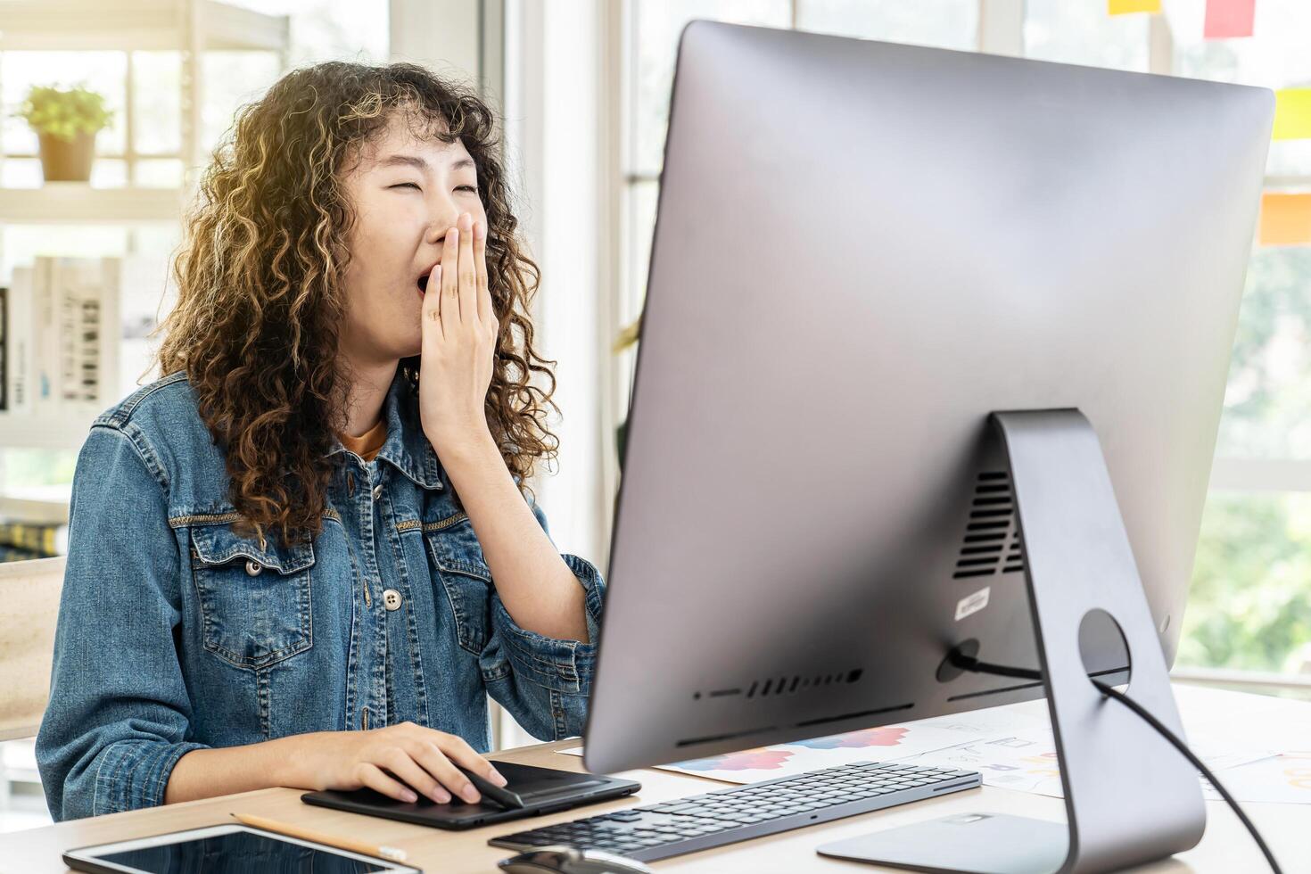 Young asian business woman yawns at office desk in front of laptop. Businesswoman covering mouth out of courtesy chain reaction drowsiness, suffer from lack of sleep photo