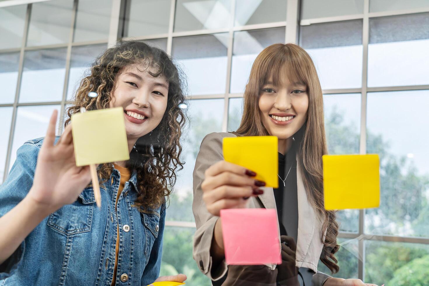 Asian creative business women are writing note on the text sheet after thinking new idea and planning project with colleague, Two employee brainstorming idea on glass wall and sticky photo