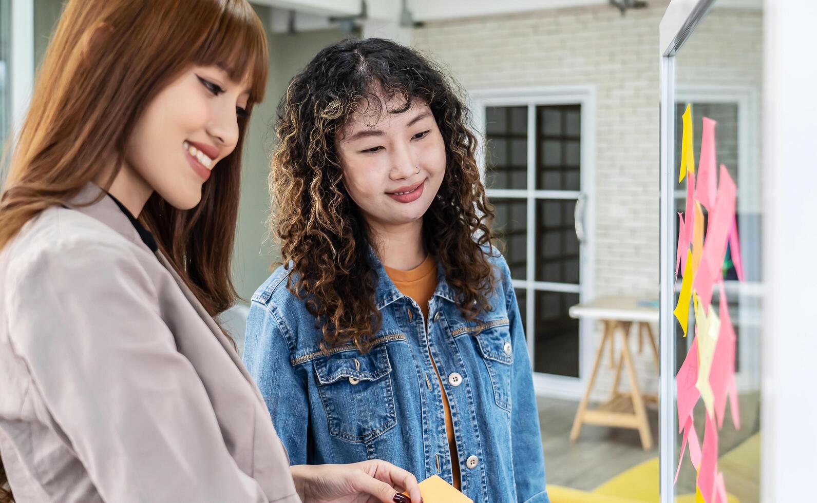 Asian creative business women are writing note on the text sheet after thinking new idea and planning project with colleague, Two employee brainstorming idea on glass wall and sticky photo