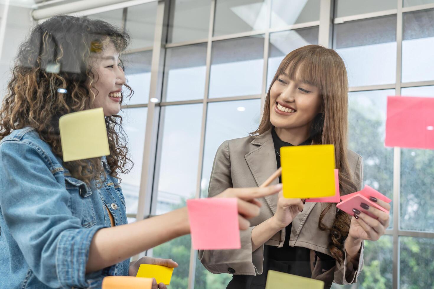 Asian creative business women are writing note on the text sheet after thinking new idea and planning project with colleague, Two employee brainstorming idea on glass wall and sticky photo