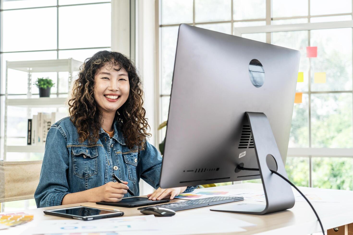 Asian young woman graphic designer smiling and looking at camera sitting at the desk with pc and tablet computer while working in office, Artist Creative Designer Illustrator Graphic Skill Concept photo
