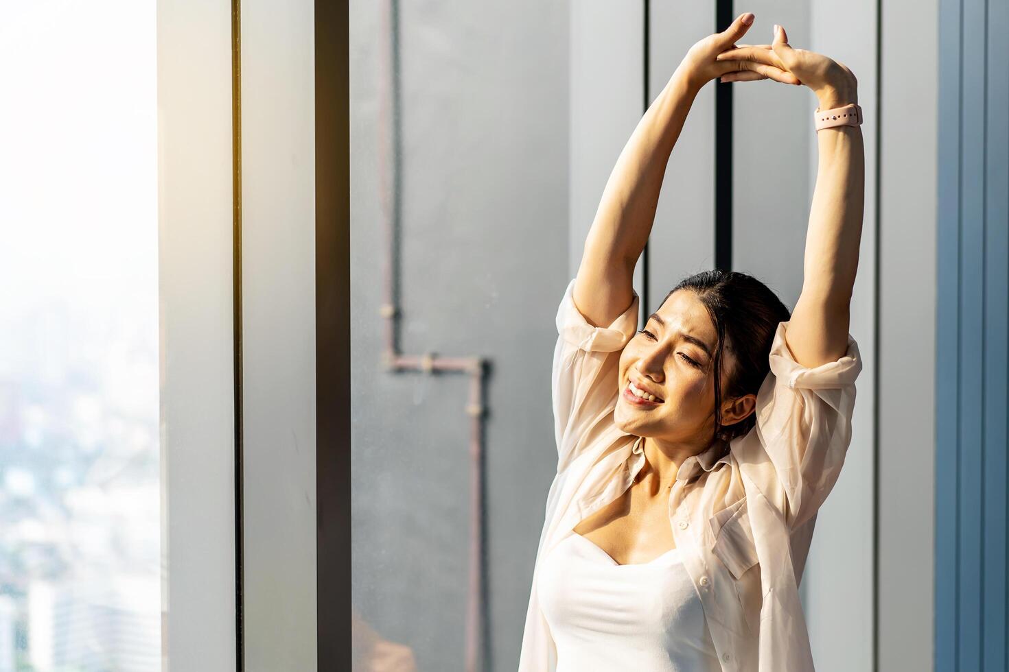 Young asian woman standing in front of big window, stretching arms and back, enjoying good morning time in office. Happy attractive millennial girl feeling no stress and peace of mind concept photo