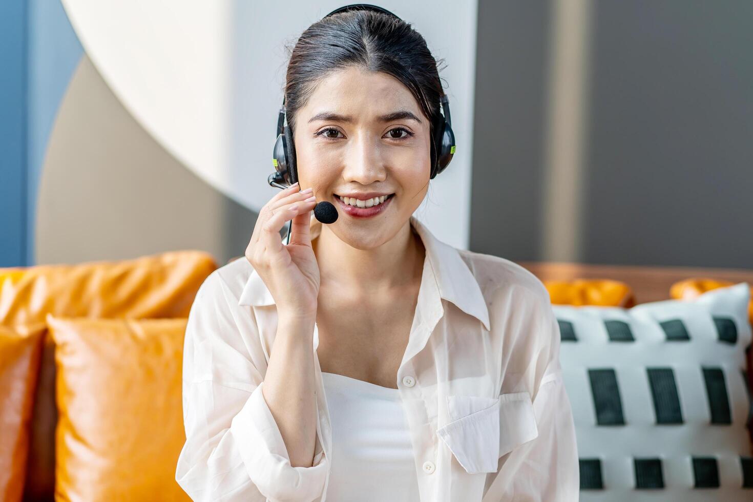 Consultant girl wearing headset with microphone sitting on sofa, freelance worker, call center or support service operator helpline. Young woman having talk with client or colleague photo
