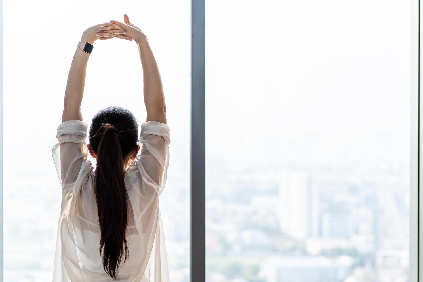 Young asian woman standing in front of big window, stretching arms and back, enjoying good morning time in office. Happy attractive millennial girl feeling no stress and peace of mind concept photo