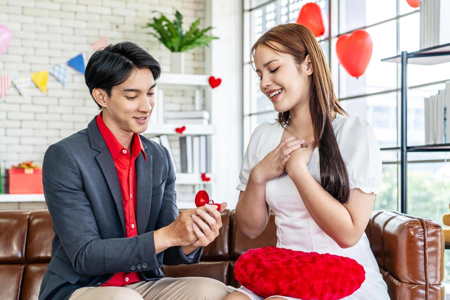 Love, relationships, proposal, Happy asian young man or boyfriend surprising his amazed pretty girlfriend with engagement ring in little red box sitting on the couch at home in the living room photo