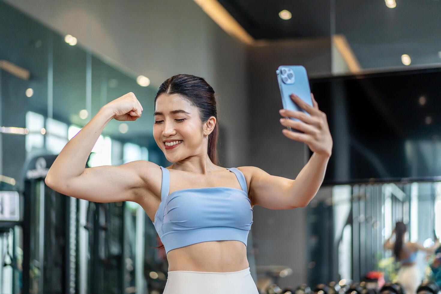 Young asian athletic woman with perfect body wearing sportswear making call with smart phone to friends or family to show her achievements and results while workout in gym, healthy lifestyle photo