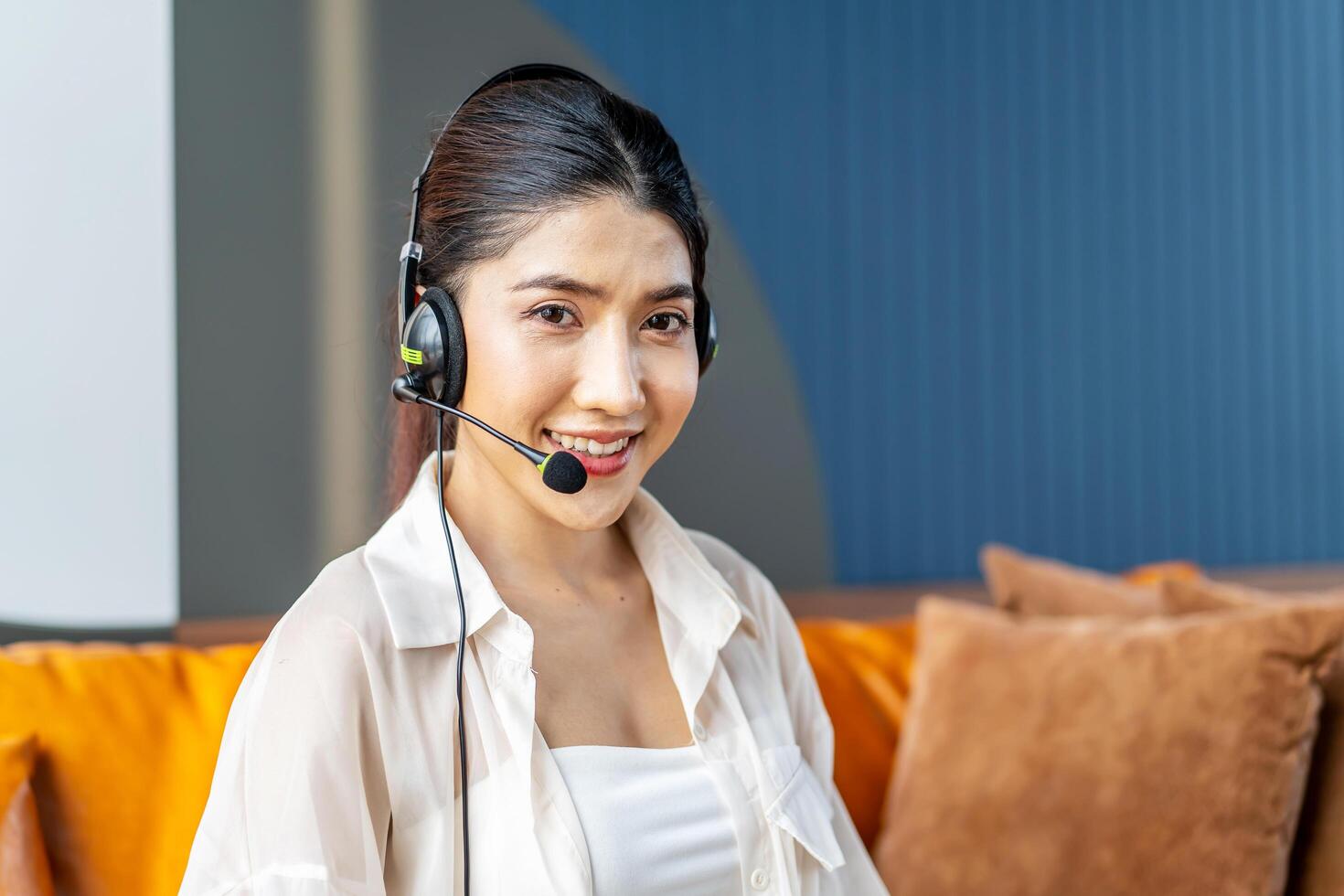 Consultant girl wearing headset with microphone sitting on sofa, freelance worker, call center or support service operator helpline. Young woman having talk with client or colleague photo