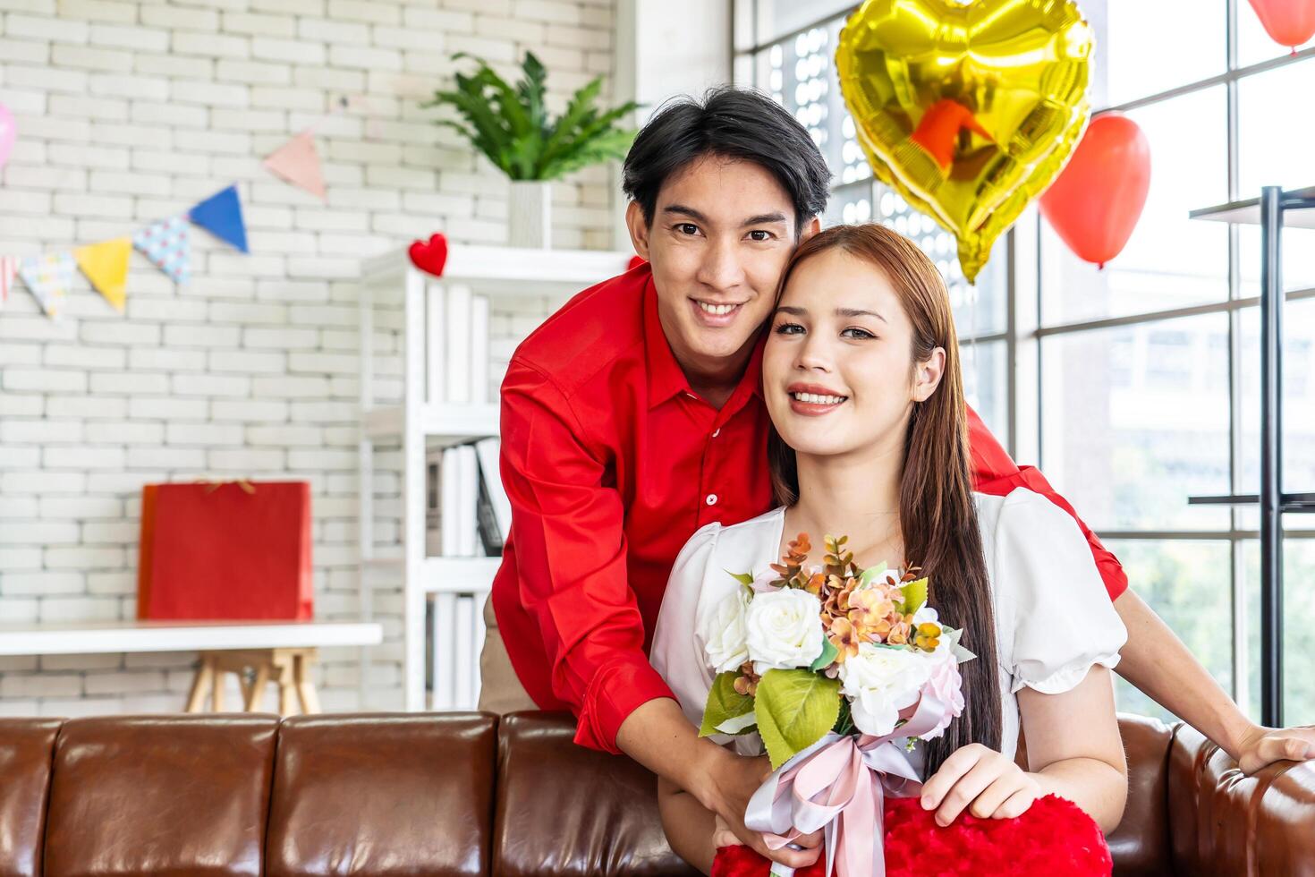 Portrait of asian young couple smiling and holding red heart pillow and bouquets in living room for romance and celebrating valentine day. Concept love story of young couple in valentine's day. photo