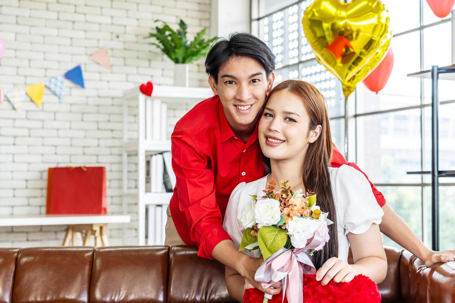 Portrait of asian young couple smiling and holding red heart pillow and bouquets in living room for romance and celebrating valentine day. Concept love story of young couple in valentine's day. photo