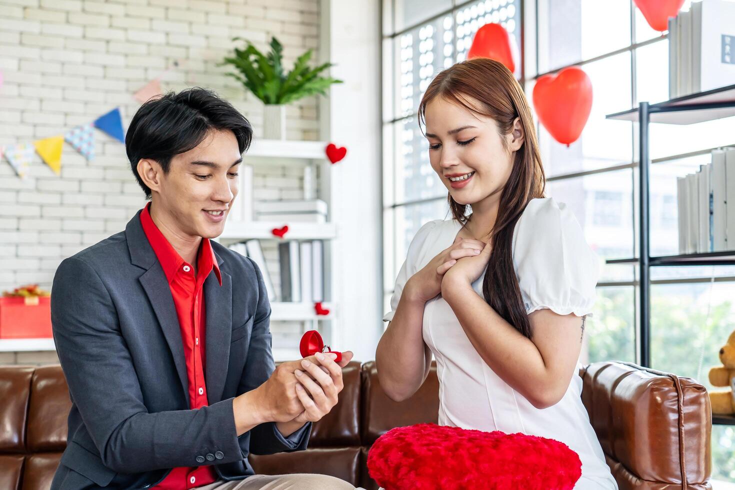 Love, relationships, proposal, Happy asian young man or boyfriend surprising his amazed pretty girlfriend with engagement ring in little red box sitting on the couch at home in the living room photo