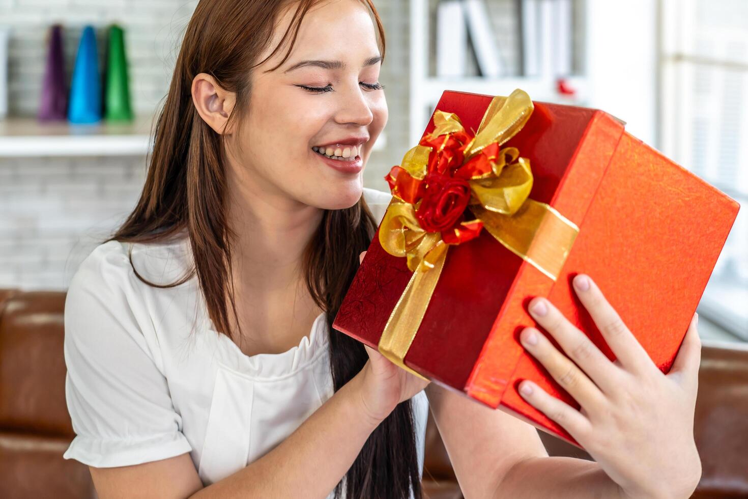 Attractive beautiful Asian woman holding a gift box while sitting on the sofa in the living room, Smiling young woman holds a Valentine's Day gift from her boyfriend, lover. Happy Valentine's Day photo