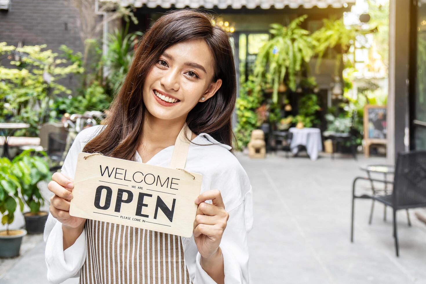 Business owner happy beautiful young asian woman in apron looking at camera, we're open sign on front door smiling welcoming clients to new cafe, People and start-up concept. photo