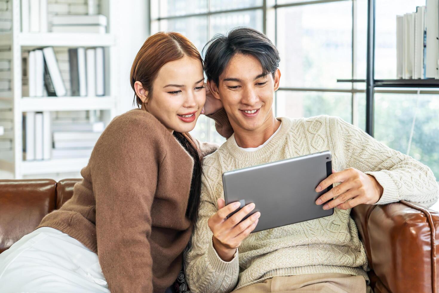 Happy asian young couple looking at digital computer tablet screen, relaxing on comfortable couch. Loving couple shopping online in internet store, watching media content online photo