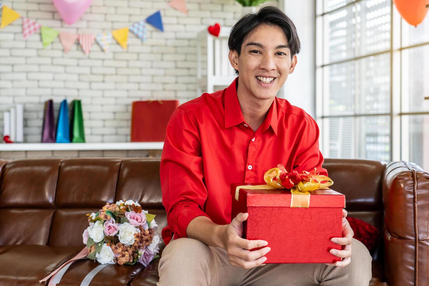 Young asian man with gift red box in his hands and bouquet, sitting on the sofa in his room on Valentine's Day, and preparing surprise for his girlfriend. Marriage proposal. Attention and care photo