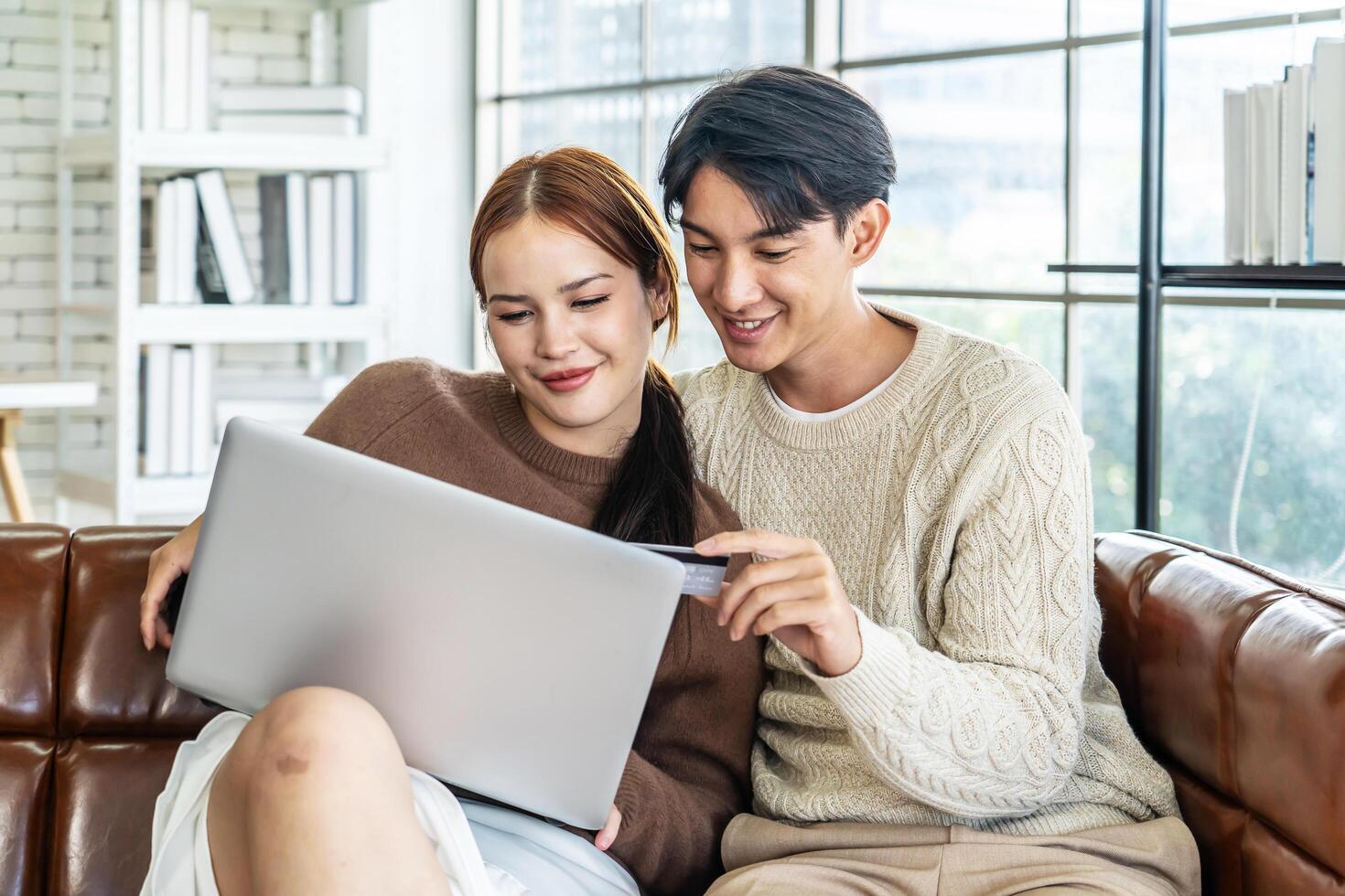 Asian Couple Use Laptop Computer, while Sitting on the sofa in living room of their Apartment. Boyfriend and Girlfriend Talk, Shop on Internet, Choose Product to Order Online, Watch Streaming Service photo