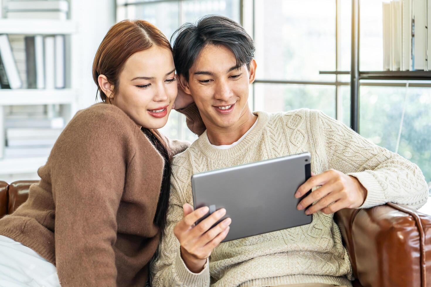 Happy asian young couple looking at digital computer tablet screen, relaxing on comfortable couch. Loving couple shopping online in internet store, watching media content online photo