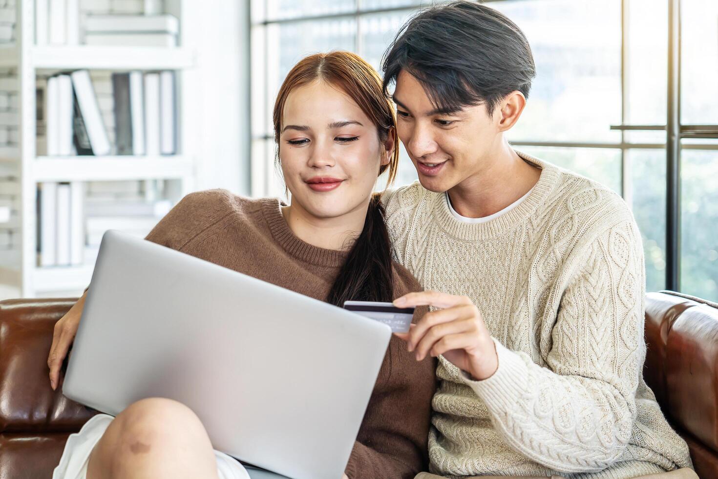 Asian Couple Use Laptop Computer, while Sitting on the sofa in living room of their Apartment. Boyfriend and Girlfriend Talk, Shop on Internet, Choose Product to Order Online, Watch Streaming Service photo