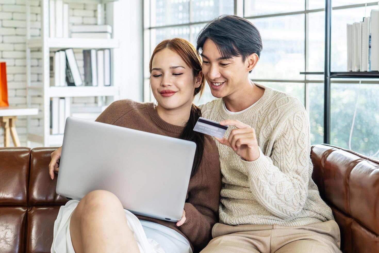 Asian Couple Use Laptop Computer, while Sitting on the sofa in living room of their Apartment. Boyfriend and Girlfriend Talk, Shop on Internet, Choose Product to Order Online, Watch Streaming Service photo