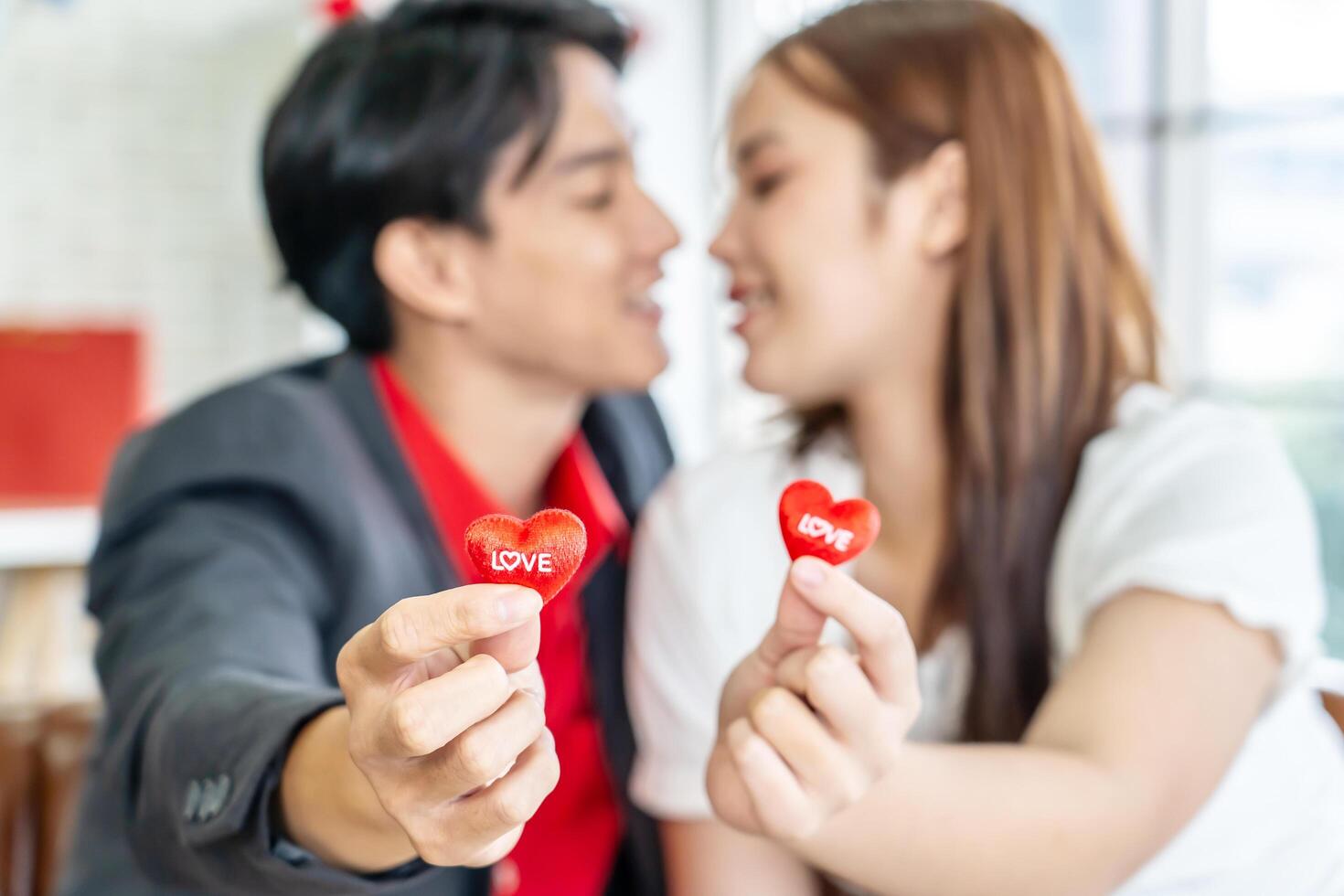 Happy Asian couple holding red hearts while sitting on sofa in living room with red heart balloons, romantic lover celebrating anniversary on Valentine Day together, selective focus photo