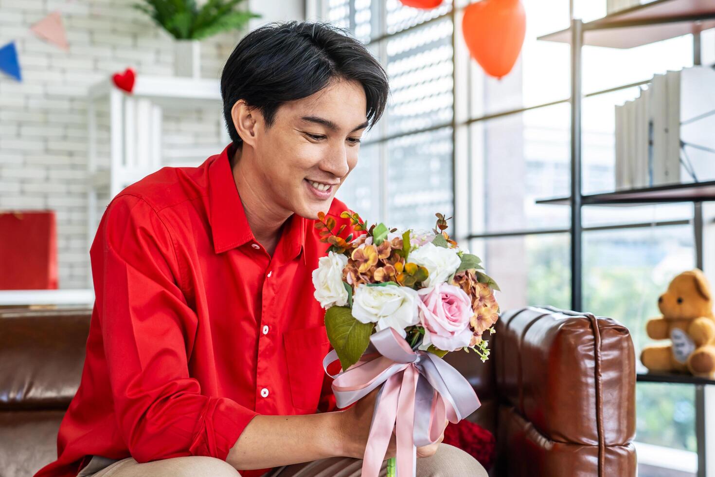 joven asiático hombre con un ramo de flores de flores en su manos, sentado en el sofá en su habitación en San Valentín día, y preparando sorpresa para su novia. preparando para fecha. atención y cuidado. foto