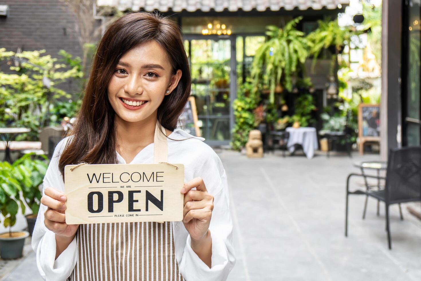 Business owner happy beautiful young asian woman in apron looking at camera, we're open sign on front door smiling welcoming clients to new cafe, People and start-up concept. photo