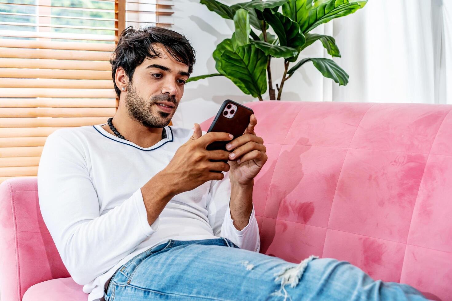 Cool gadget and app. Young smiling guy holding mobile phone, typing sms message, resting on sofa in living room, Texting And Browsing Internet, surfing web, using app, Networking In Social Media photo