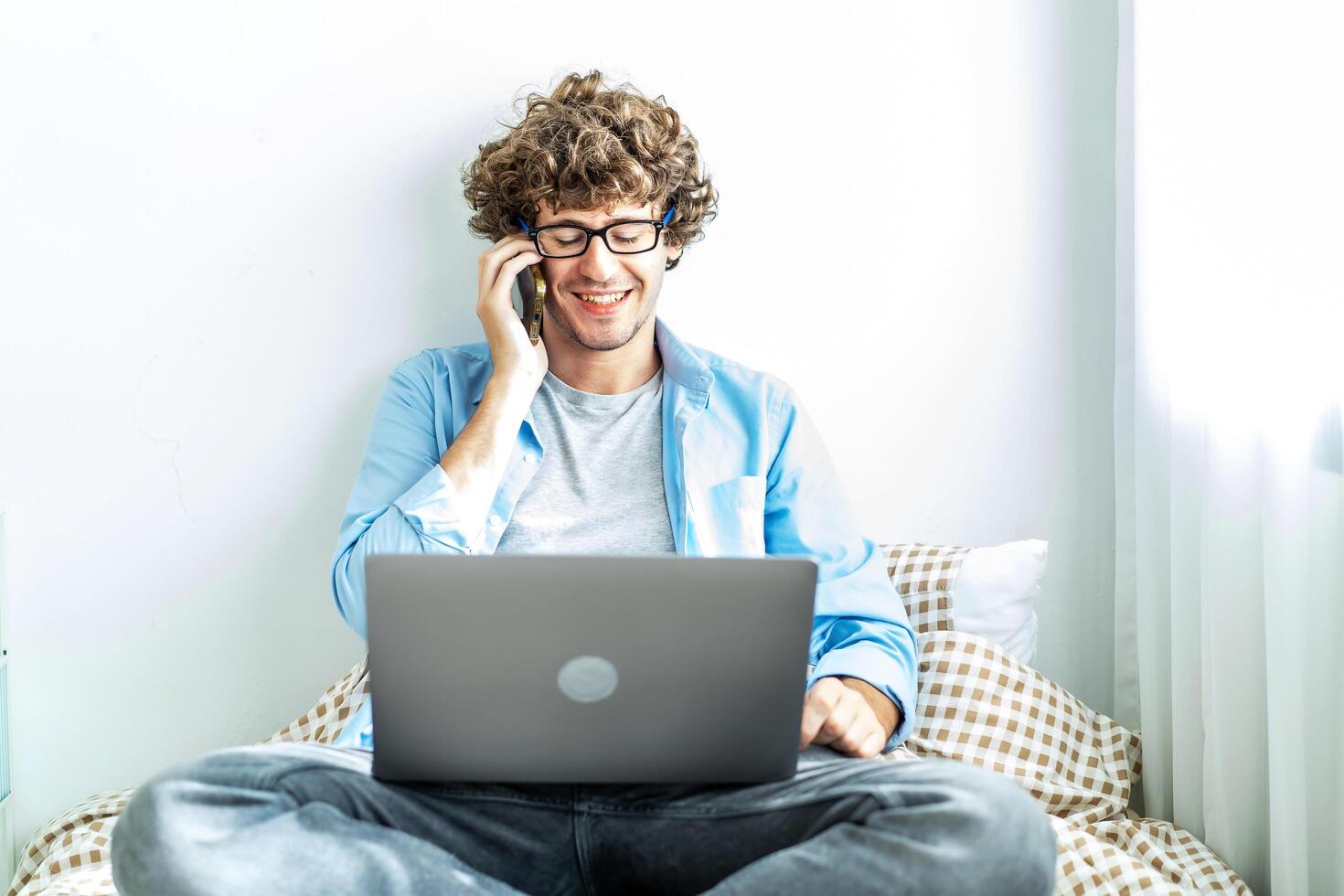 Caucasian Freelancer Guy Talking On Phone Using Laptop Computer Communicating Online Working on the bed in the room, Distance Work And Mobile Communication Concept. photo