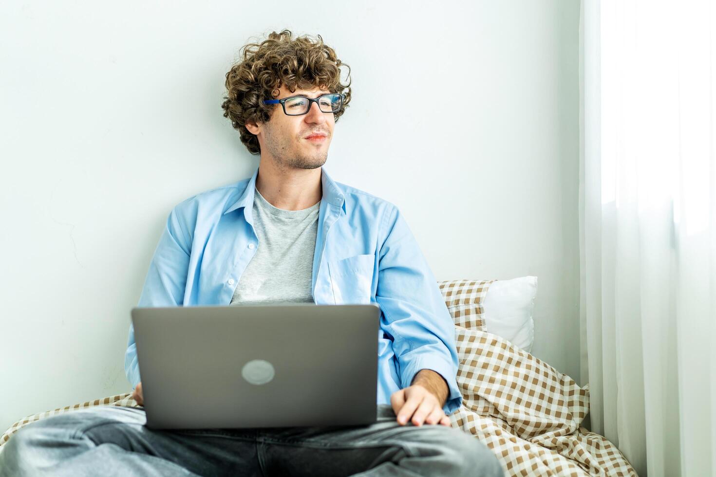 Work from home. Young caucasian man working on computer laptop and looking away, study online listening learning education course conference calling sit laying down on bed, freelancer at home photo