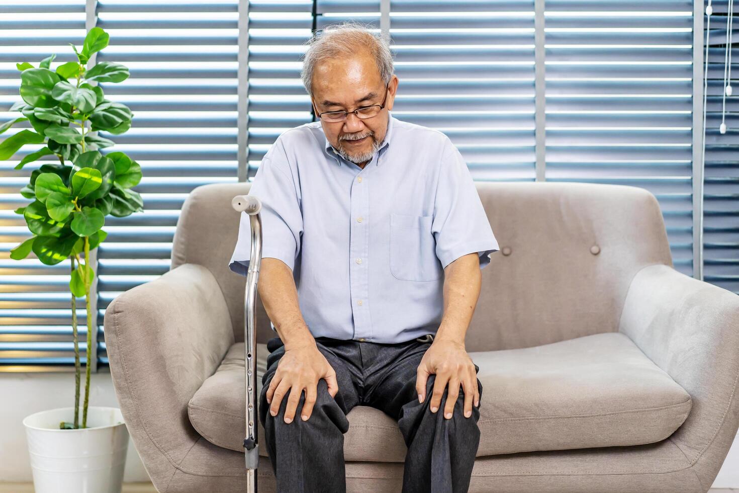 Pensive senior man leaning on walking stick while sitting on sofa. Lonely elderly man sitting in living room at home. lost in sad thoughts feels lonely, recollect life moments photo