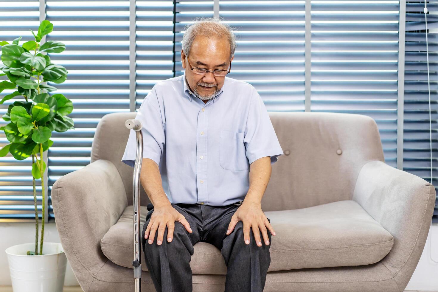 Pensive senior man leaning on walking stick while sitting on sofa. Lonely elderly man sitting in living room at home. lost in sad thoughts feels lonely, recollect life moments photo