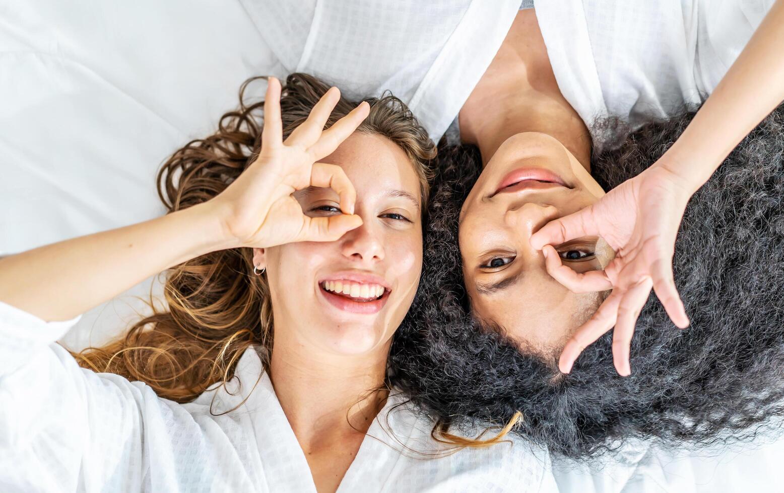 Top view self portrait of caucasian and black women wearing sleep with happy face smiling doing ok sign with hand on eye looking through fingers, looking at camera photo