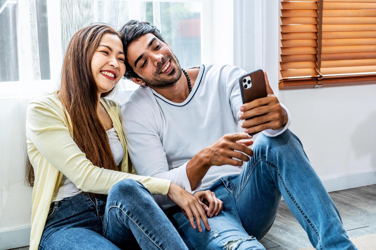 Relaxed young interracial couple or friends using a generic mobile phone together sitting in the living room at home, Happy young man and woman with smartphone photo