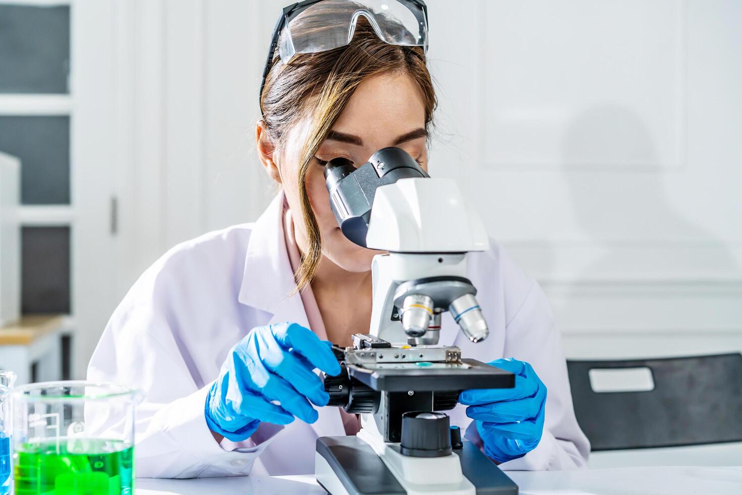 Smiling young asian woman wearing scientist uniform working at scientist laboratory with looking through a microscope in a laboratory. Young scientist doing some research. photo