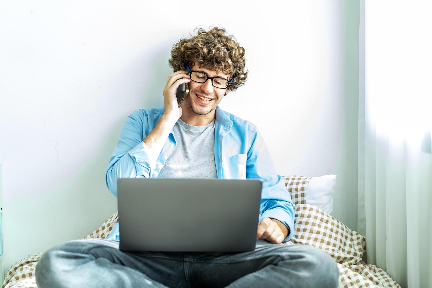 Caucasian Freelancer Guy Talking On Phone Using Laptop Computer Communicating Online Working on the bed in the room, Distance Work And Mobile Communication Concept. photo