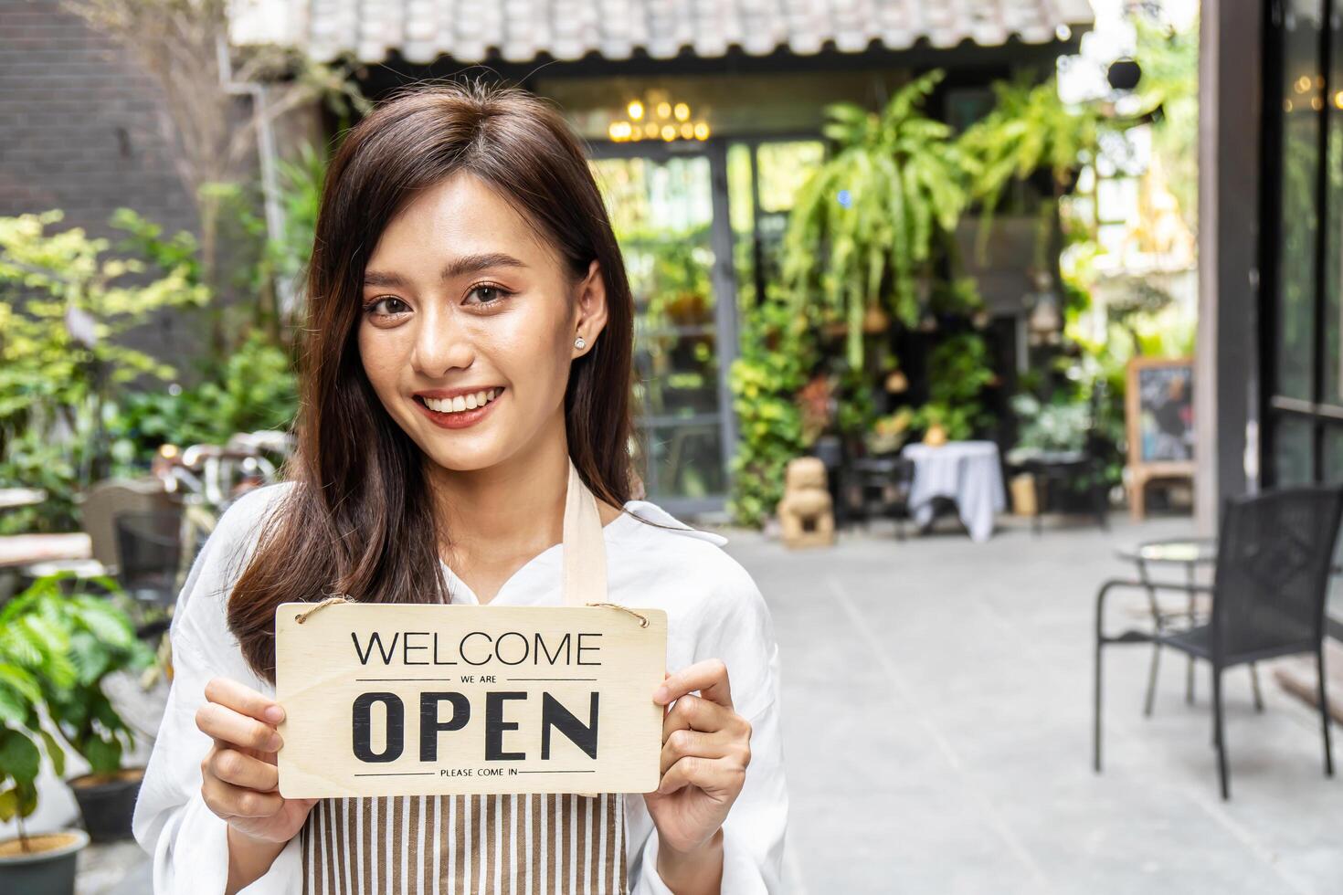 Business owner happy beautiful young asian woman in apron looking at camera, we're open sign on front door smiling welcoming clients to new cafe, People and start-up concept. photo