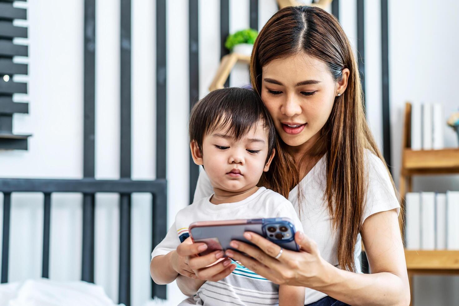 Young asian mother her little adorable son using mobile phone looking at screen enjoying watching funny social media doing online shopping relaxing on bed at home together photo