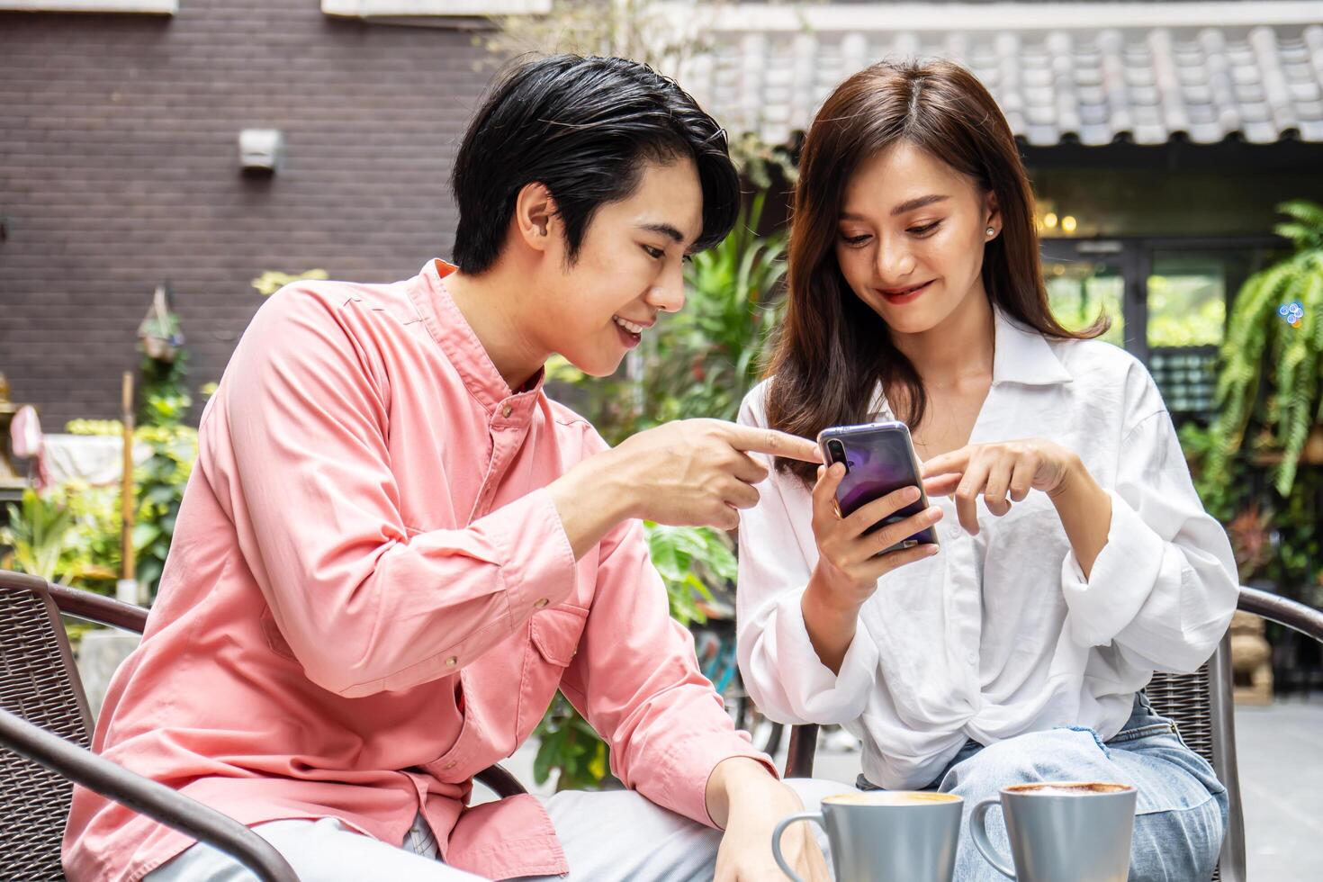 Happy Young asian woman sitting with boyfriend at cafe table, discussing over on smartphone, Young couple looking at mobile phone shopping online and share information together in coffee shop photo