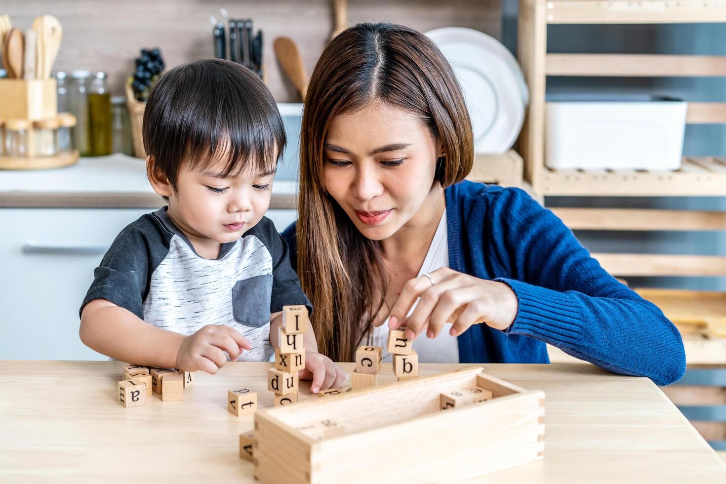 Young asian mother and little son enjoy to play wooden jigsaw puzzle together in area of kitchen of their house and they look happiness, childhood and motherhood concept photo
