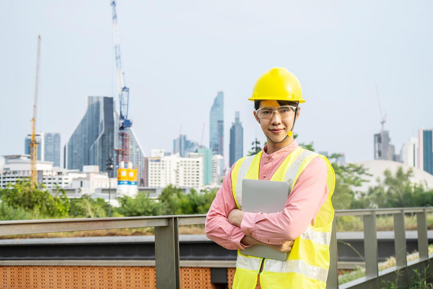 Engineer or construction worker Asian man is holding the laptop computer and crossed arms pose on the top of the roof at building, Hard hat help protect with operation photo
