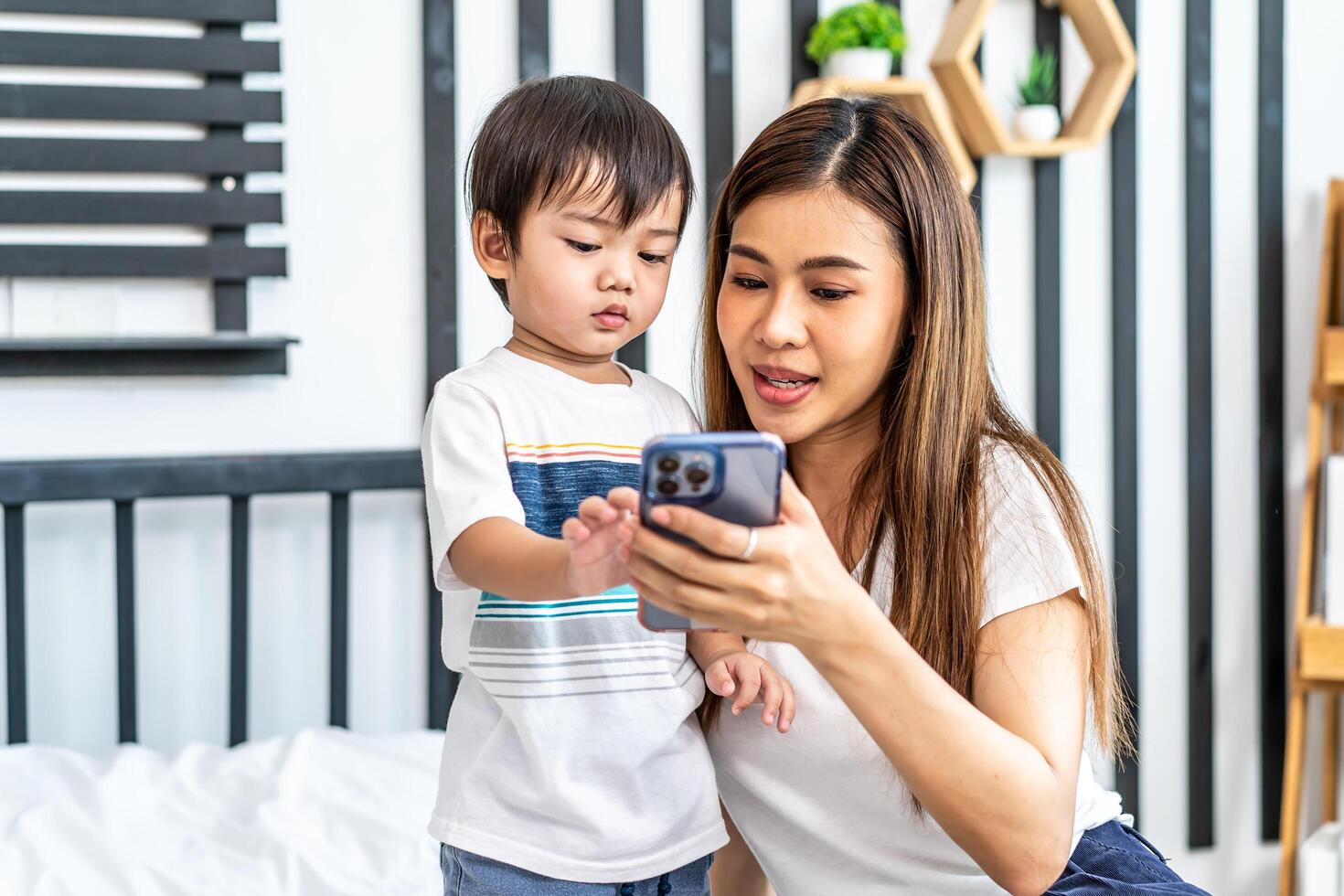 Young asian mother her little adorable son using mobile phone looking at screen enjoying watching funny social media doing online shopping relaxing on bed at home together photo