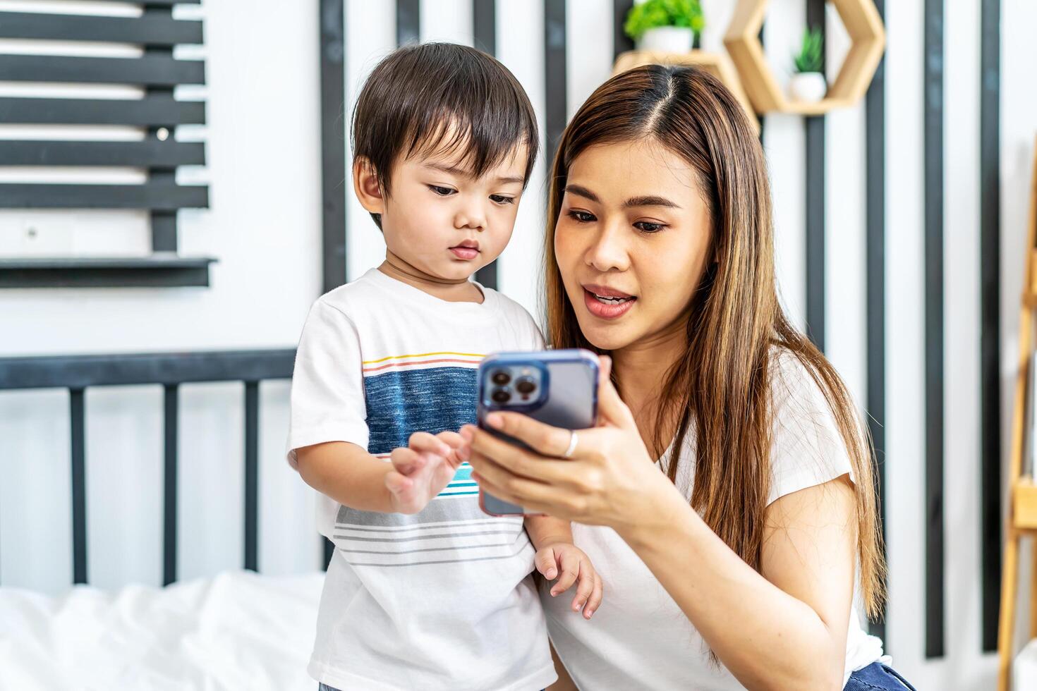 Young asian mother her little adorable son using mobile phone looking at screen enjoying watching funny social media doing online shopping relaxing on bed at home together photo