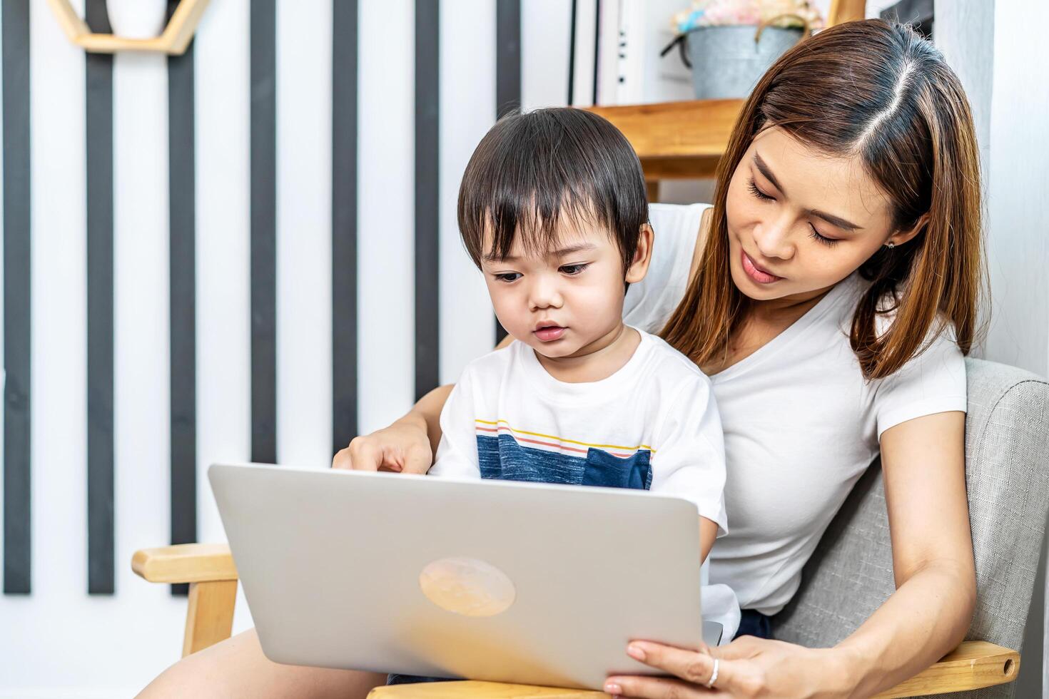 Young asian mother her little adorable son using laptop looking at computer screen enjoying watching funny social media doing online shopping relaxing on sofa at home together photo