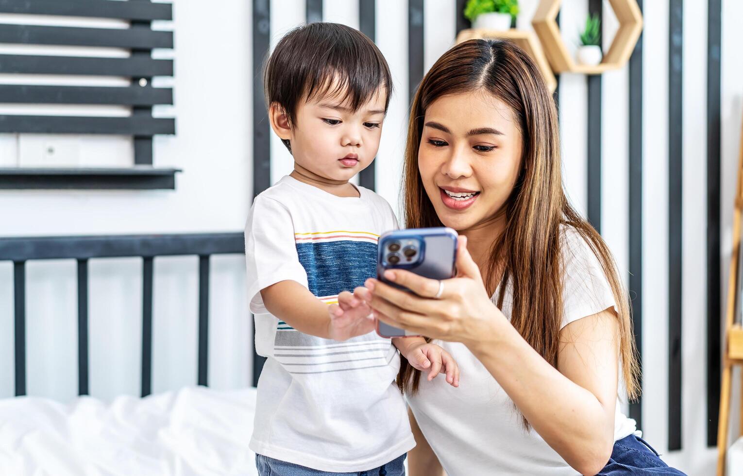 Young asian mother her little adorable son using mobile phone looking at screen enjoying watching funny social media doing online shopping relaxing on bed at home together photo