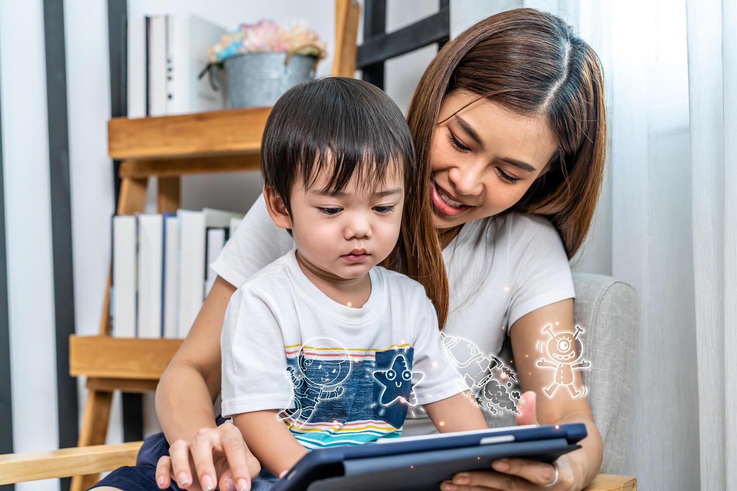Young asian mother her little adorable son using tablet looking at screen enjoying watching funny cartoon social media online relaxing on sofa together, Media icons flying over the tablet photo