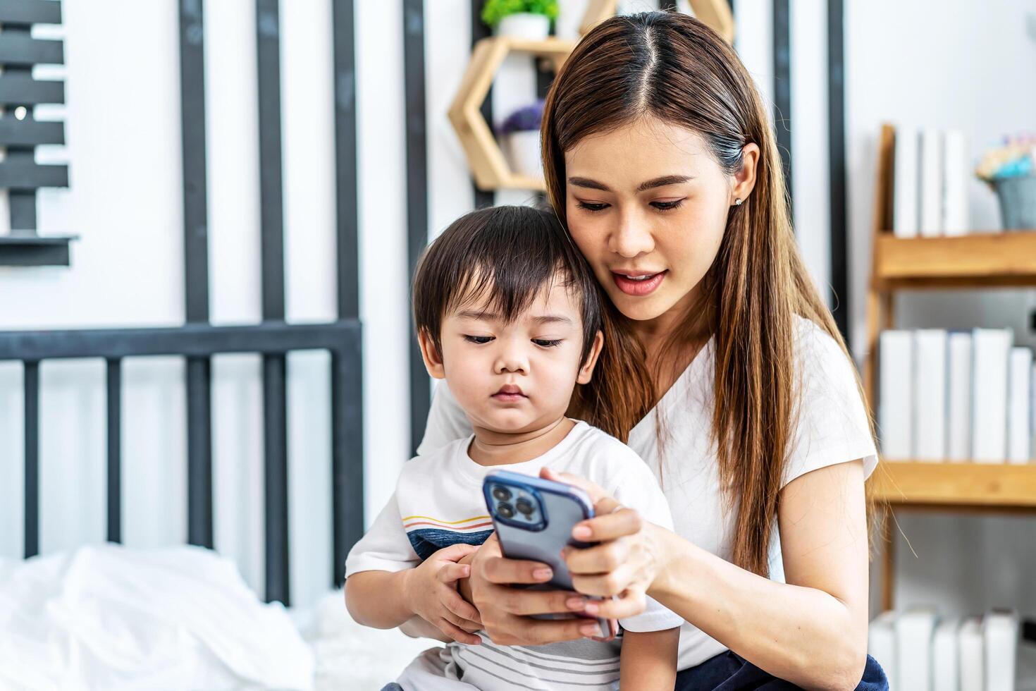 Young asian mother her little adorable son using mobile phone looking at screen enjoying watching funny social media doing online shopping relaxing on bed at home together photo