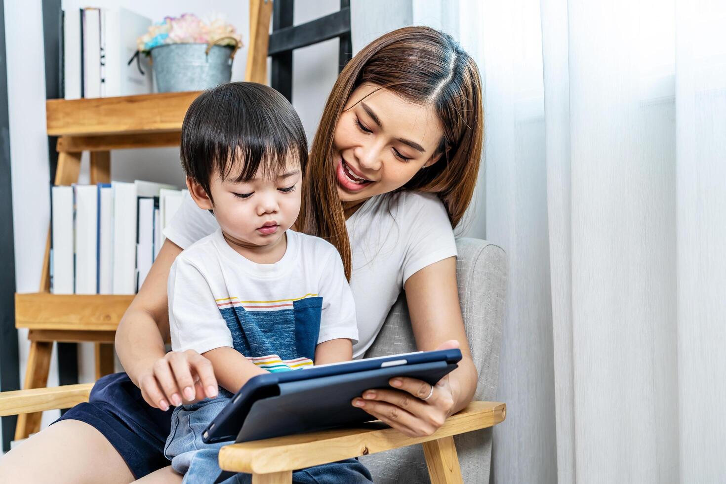Young asian mother her little adorable son using tablet looking at screen enjoying watching funny social media doing online shopping relaxing on sofa at home together photo