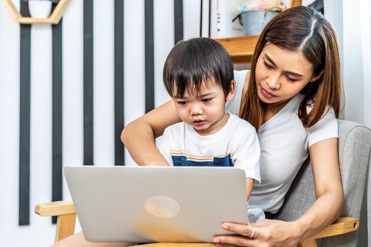 Young asian mother her little adorable son using laptop looking at computer screen enjoying watching funny social media doing online shopping relaxing on sofa at home together photo