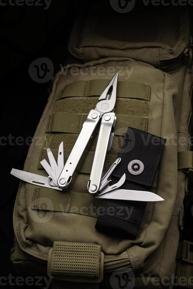 Modern multitool with many tools. A portable multitasking tool lie on a khaki-coloured military backpack. Dark back. photo