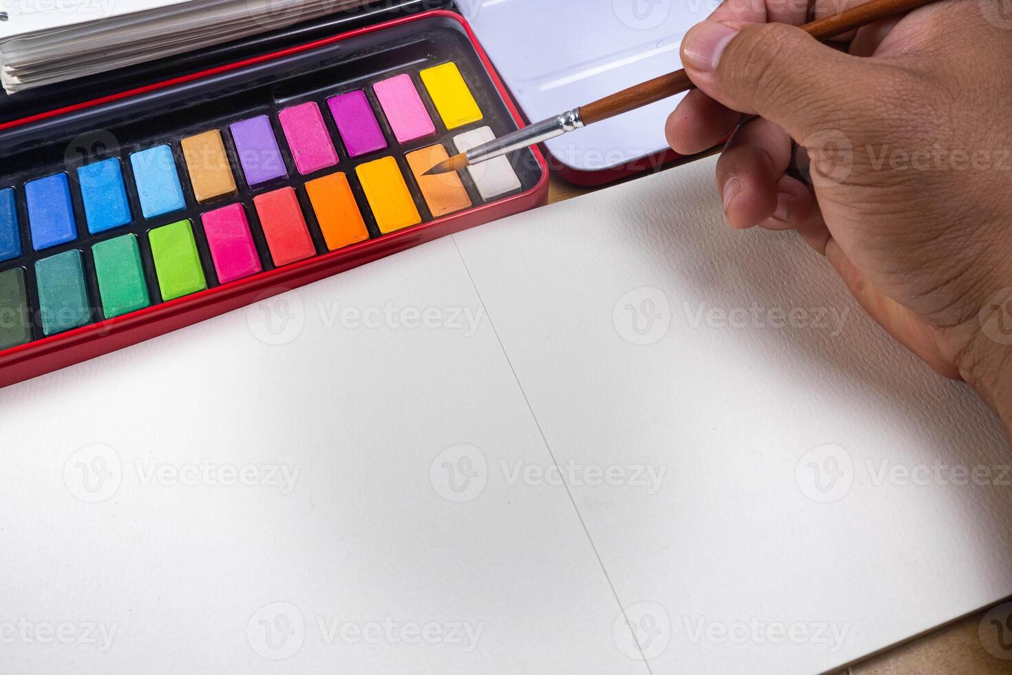 Paint accessories and watercolor and blank book on wooden table. Someone painting in blank drawing book with brush and watercolor color palette. Flat lay, top view. photo